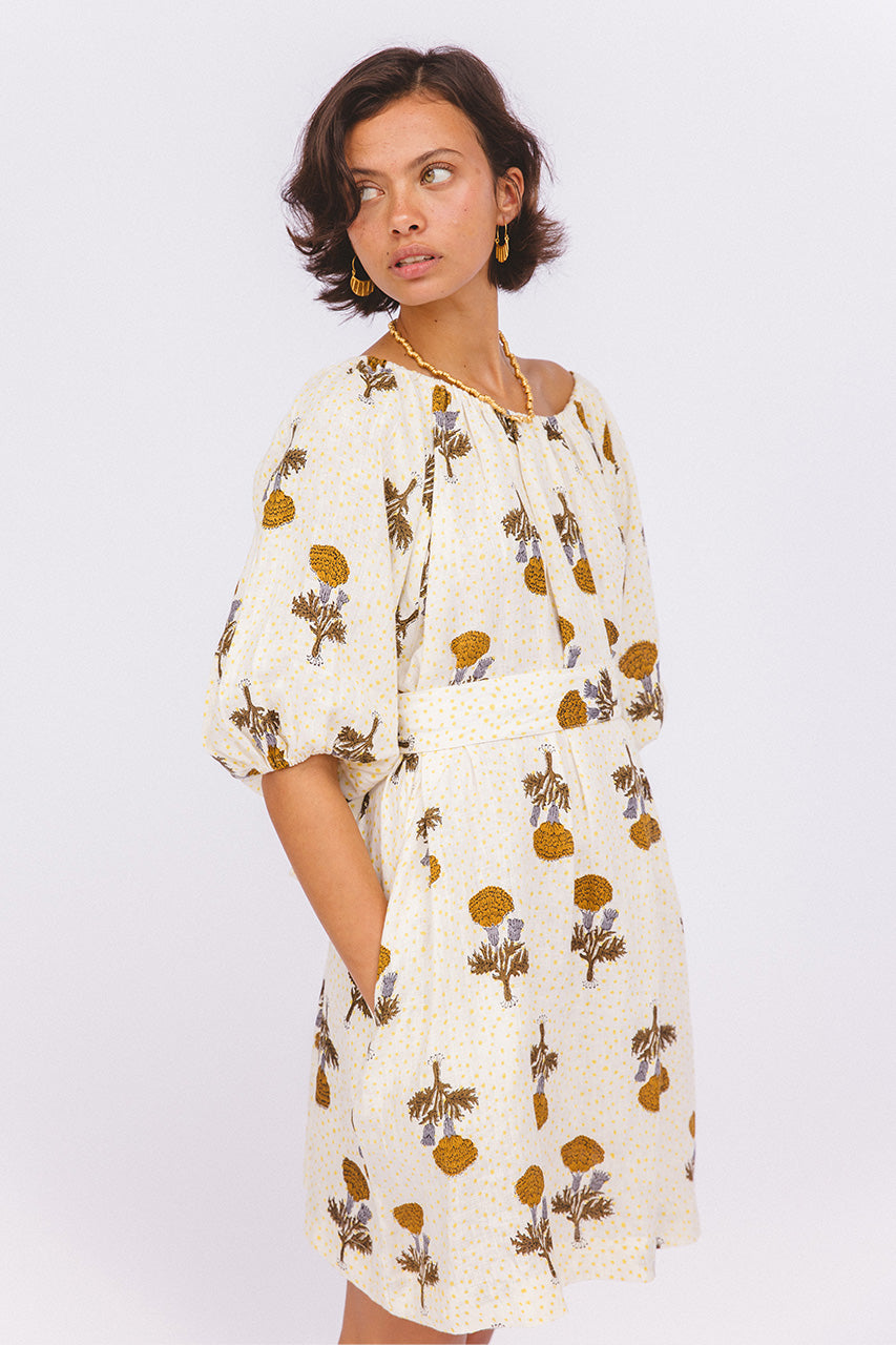 Dahlia Mini Dress Marigold — studio model side glance with hand in pocket, on-shoulder tie neckline, white linen with golden marigold flower hand block print, Jaipur artisan made mini dress | Daughters of India