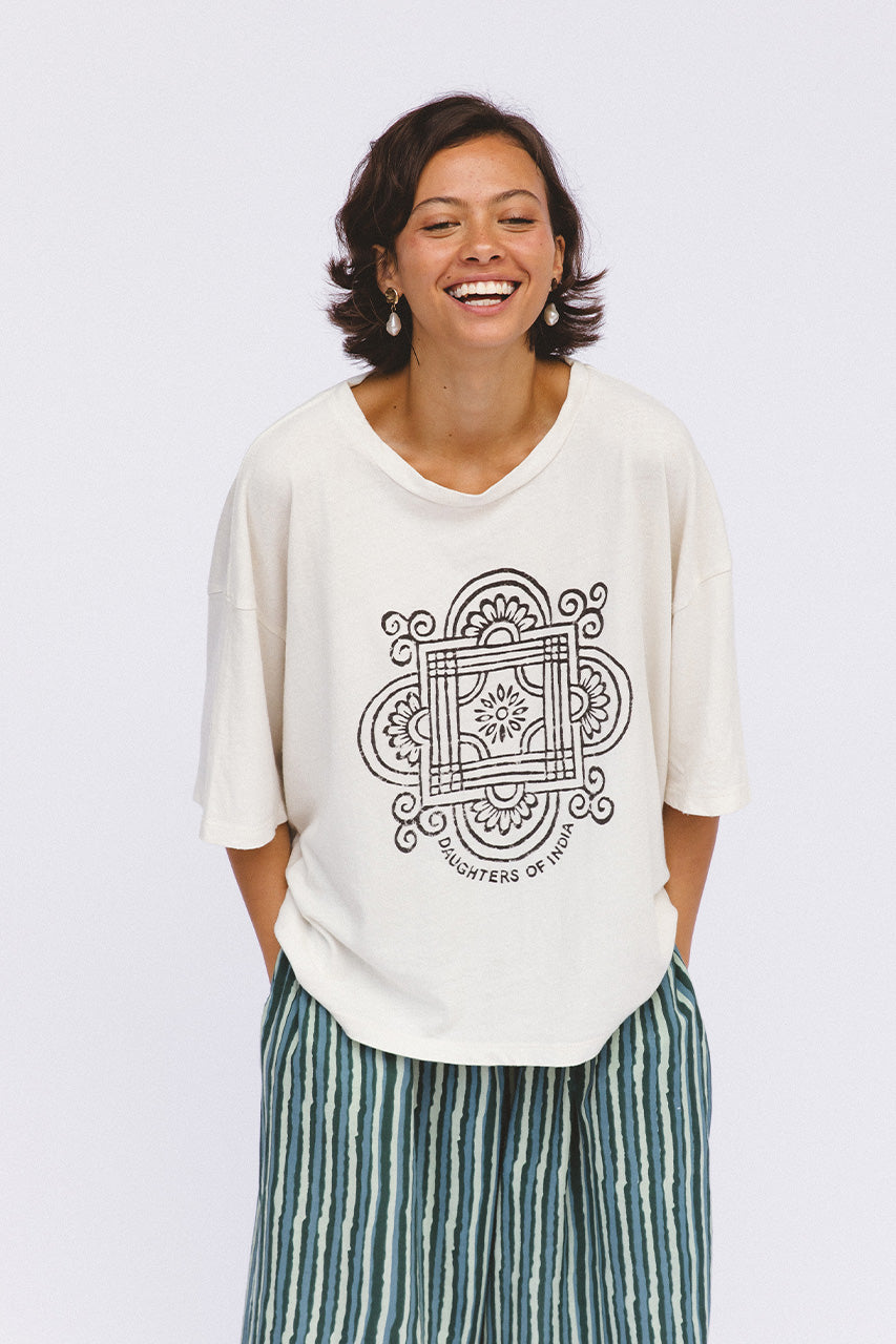 Shanti Tee White — studio front view smiling, white oversized cotton tee with charcoal hand-printed mandala logo, paired with spearmint striped pants, white backdrop | Daughters of India