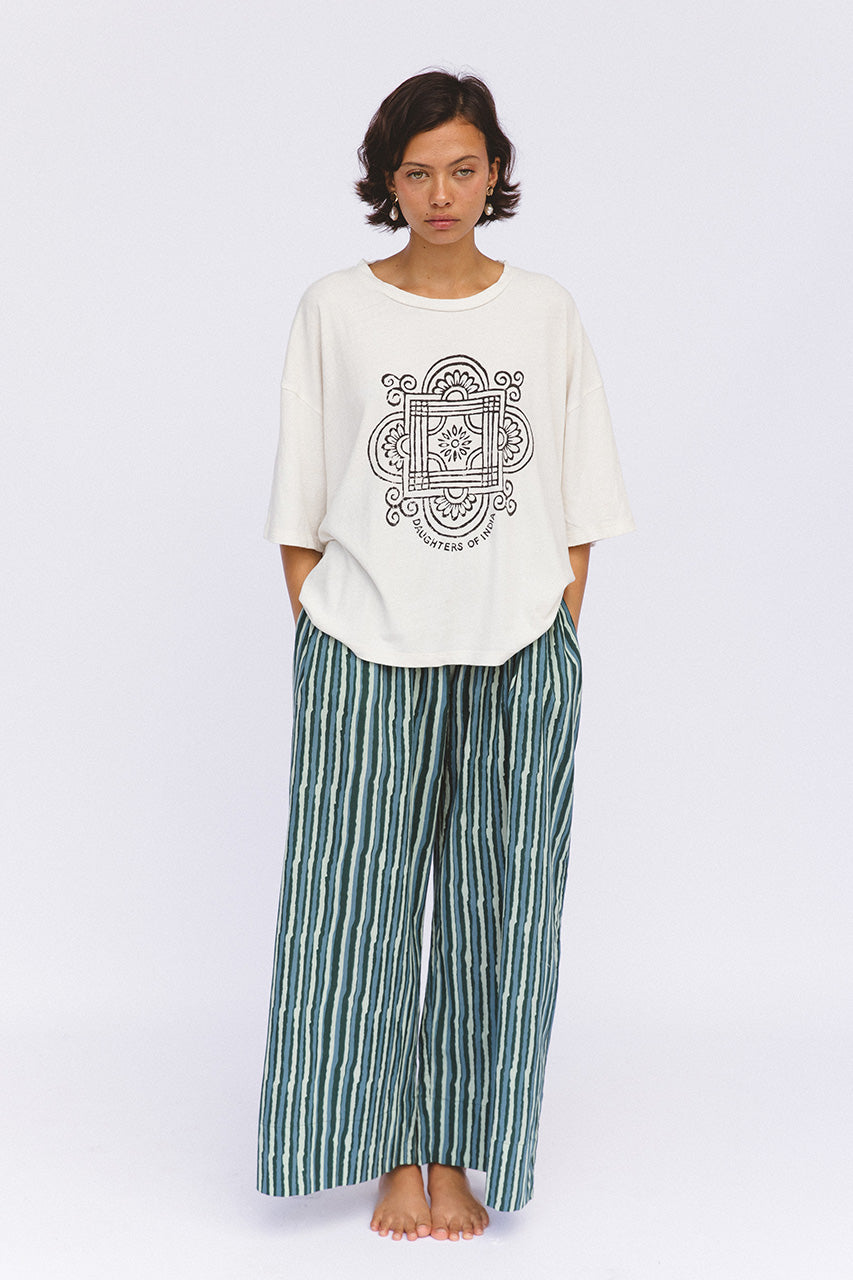 Shanti Tee White — studio front view with hands in pockets, white oversized cotton tee with charcoal mandala logo, paired with spearmint striped pants, white backdrop | Daughters of India