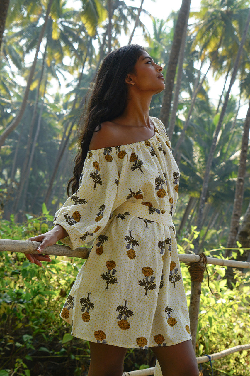 Dahlia Mini Dress Marigold — model in tropical palm forest setting, off-shoulder styling, looking up, hand block print linen mini with golden marigold botanical motif, ethical boho travel dress | Daughters of India